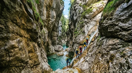 Vista grandangolare di un gruppo di persone in muta da canyoning che affrontano una cascata alpina tra le gole rocciose, con equipaggiamento di sicurezza visibile