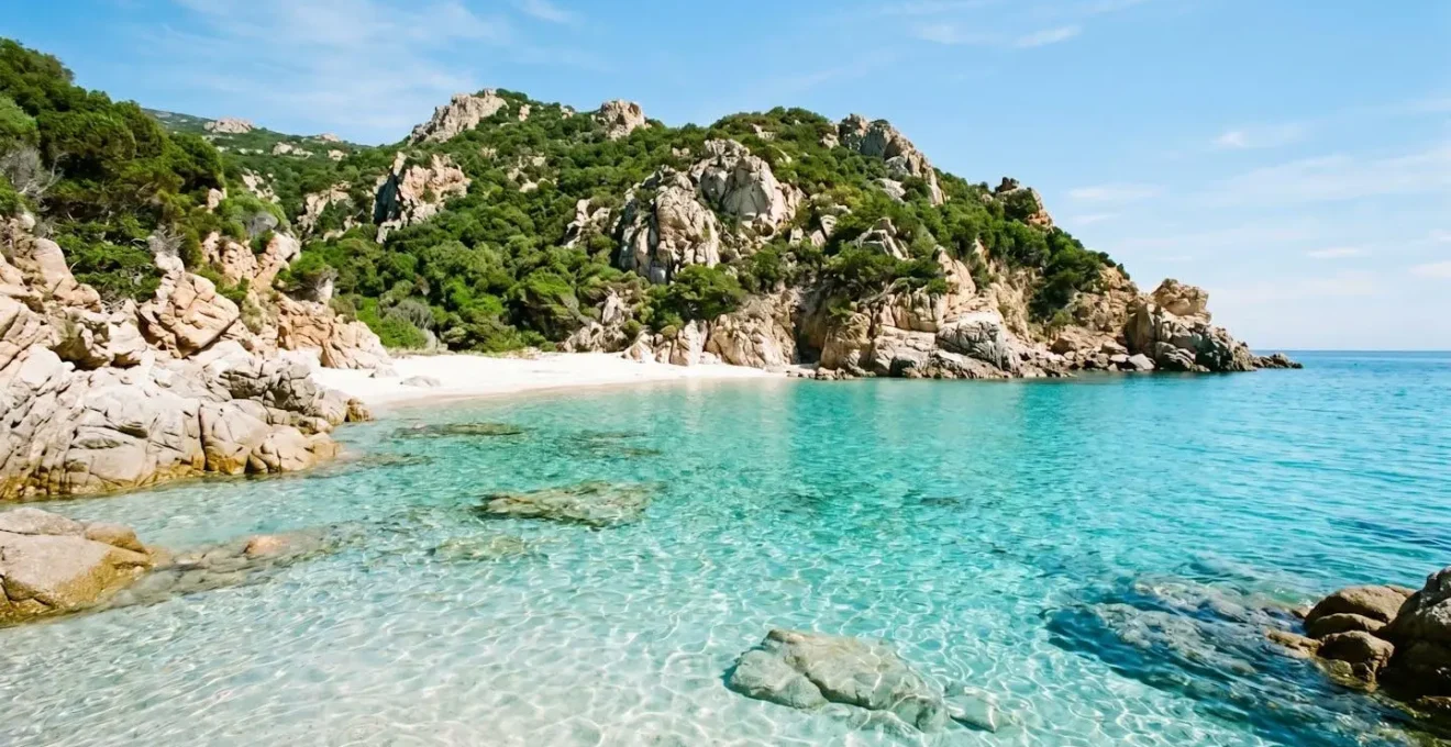 Vista dal mare di una piccola baia corsa con acqua turchese trasparente, spiaggia di sabbia bianca orlata da rocce granitiche chiare e vegetazione mediterranea verde brillante, sotto un cielo azzurro intenso