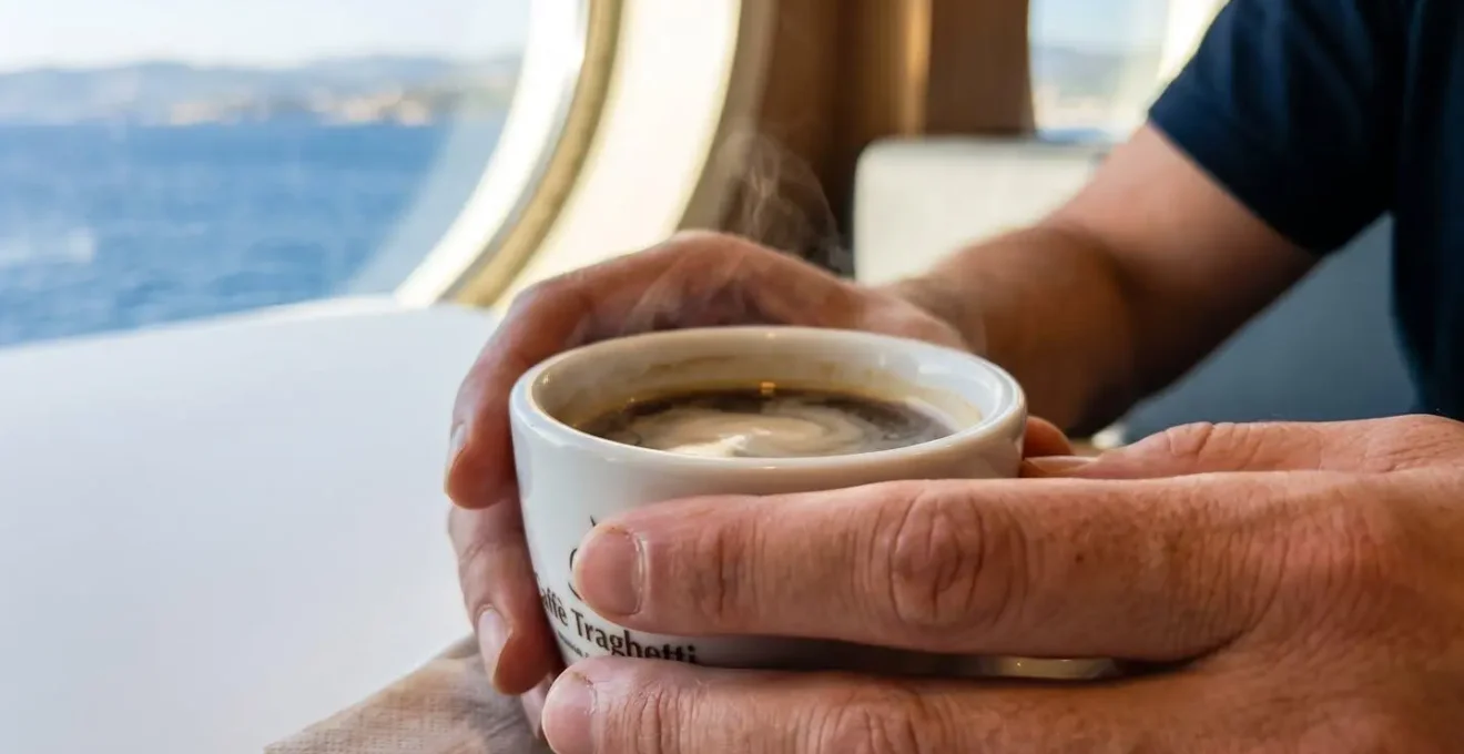 Primo piano di mani che tengono una tazza di caffè fumante su un tavolino moderno di traghetto, con uno oblò sullo sfondo sfocato che mostra il mare azzurro