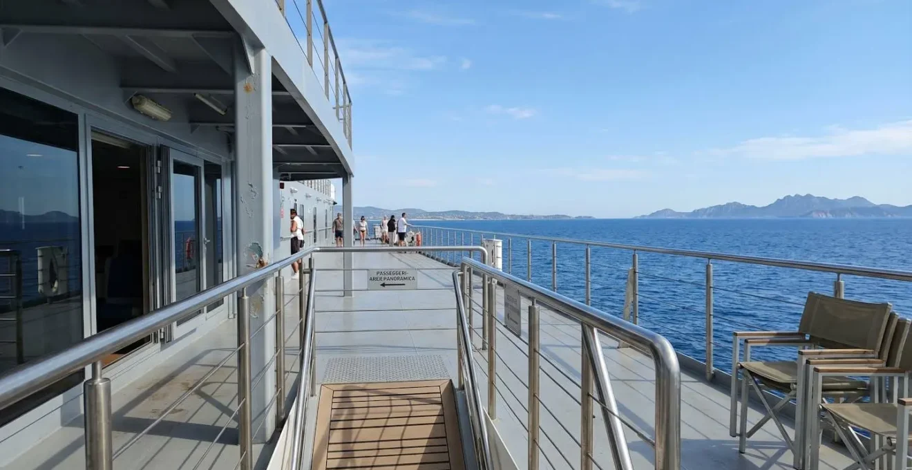 Ponte esterno panoramico di un traghetto moderno con ringhiere metalliche contemporanee, vista sul mare Mediterraneo blu intenso sotto un cielo sereno, ambiente spazioso e pulito